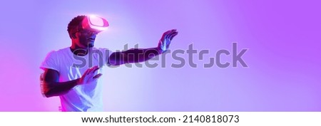 Excited young African American guy using VR glasses, touching something invisible at copy space, millennial black man experiencing virtual reality adventure on neon studio background, panorama banner