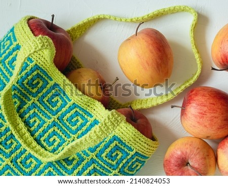 Modern blue yellow crochet bag for shopping with fresh red apples on a white background. Colored crochet bag with ethnic geometric pattern. Sustainable, reusable bag.