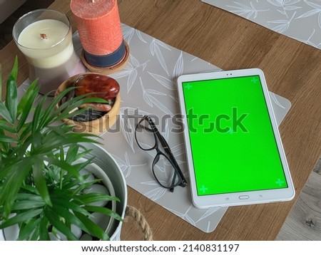 White tablet with green screen laying on wooden table next to candle, natural stones and green plant