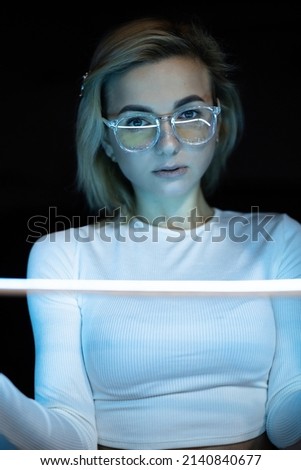 The portrait of beautiful woman in white clothes, top, shiny shimmer makeup and stars with blue led stripes, neon light on black background. Futuristic style girl in glasses, sadly emotions. Future.