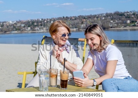 Mother and daughter spend quality time by the river together, bonding, drinking coffee, and looking at photos on their cell phone.