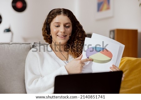 Beautiful ambitious businesswoman in a white sweater sitting in the living room in headphones in front of a laptop monitor. A woman holds documents, graphics in her hand, makes a video call, smiles