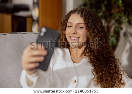 A young woman who loves to take selfies, sitting at home on a gray sofa, holding a phone in his right hand, happy with the result, she smiles at the smartphone screen