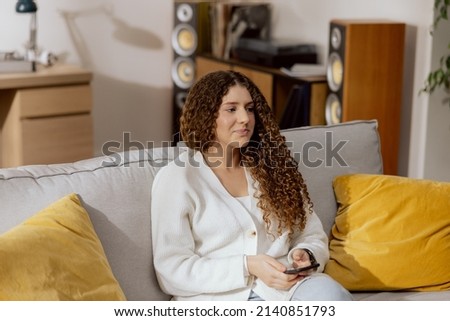 Contented woman sits on the sofa in the living room holding phone in her hands, a girl sends messages to friends, writes a blog post, replies to emails, chats, taps on the smartphone screen