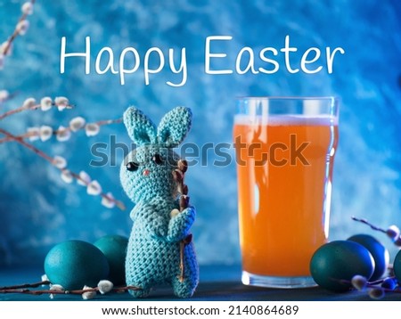 Happy Easter creative greeting. Crocheted Easter bunny on a blue background and a glass of craft beer. Willow branch and painted eggs