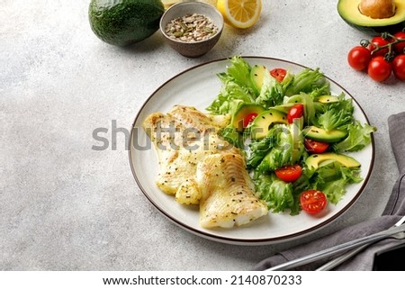Fried cod fillet with salad, fresh green leaves, avocado, seeds, tomatoes cherry. Grey background. Copy space
