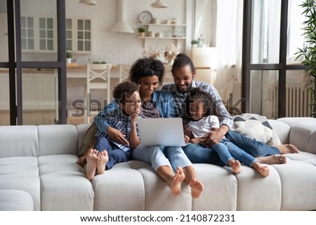 Happy African American family enjoying leisure time at laptop, resting on couch, watching movie together. Millennial couple of parents and sibling kids relaxing at home, making video call