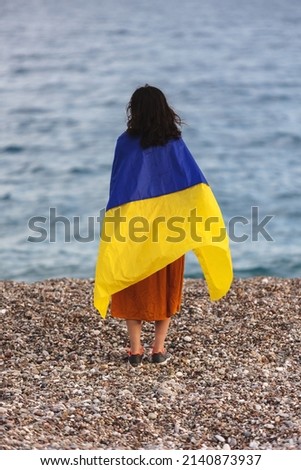 A girl stands by the sea with a Ukrainian flag, support for Ukraine, a sad Ukrainian woman, the flag flutters in the wind
