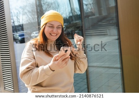 Portrait of a satisfied young woman holding mobile phone and celebrating outdoors. Happy girl walking on city street, hipster female in yellow hat overjoyed with winning online contest on web site Royalty-Free Stock Photo #2140876521