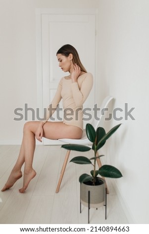 Full-lenght profile portrait of slim adorable lady in beige body is siting in stylish white room and touching her hair. High quality photo