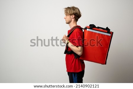 Young courier in a red uniform t-shirt and with red food thermo bag on a shoulder standing isolated on white background studio. Food delivery service.
