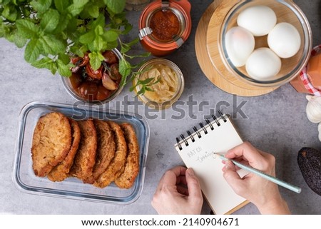 Overhead view of a woman hand writing in a spiral notebook BATCH cooking next to some glass jars full of meal. Royalty-Free Stock Photo #2140904671