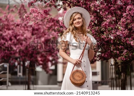 Fashionable elegant happy smiling woman wearing trendy spring, summer white dress, hat, with small shoulder round wicker bag, posing near blooming trees. Copy, empty space for text Royalty-Free Stock Photo #2140913245
