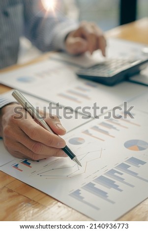 Businessman holding pen pointing to marketing graph and working on the laptop computer, working online and analyzing vertical picture financial documents.
