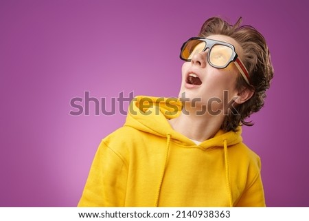 Portrait of a funny surprised teen boy in bright yellow hoodie looking up with surprise and excitement. Bright purple background with copy space. Education. Youth lifestyle, success. Emotions. Royalty-Free Stock Photo #2140938363