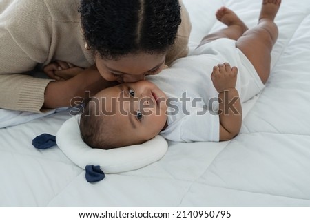 Little cute African American newborn baby lying on bed and looking smiling while young mother is kissing at infant cheek with love and tenderness at home. Motherhood, child care concept