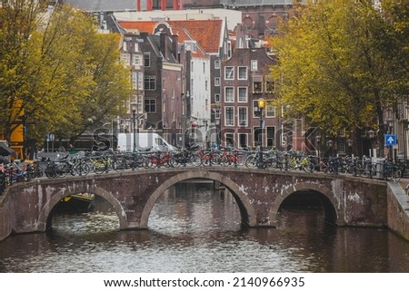 One of the many bridges in amstedam, covered with bicycles. A lot of people houses and bikes on the picture, also a picturesque trees.