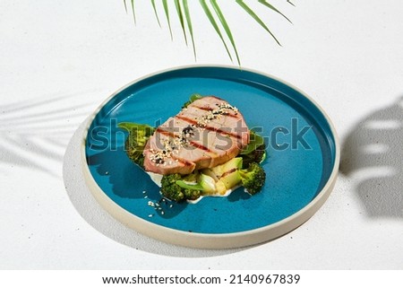 Grilled tuna steak with broccoli and zucchini in modern ceramic plate. Healthy food - roasted tuna with green vegetables. Fish dish in minimal style. Tuna fillet in blue plate with hard shadow Royalty-Free Stock Photo #2140967839