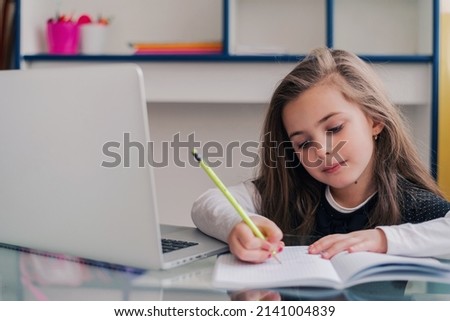 A happy kid student writing in a notebook while learning for school at the home desk.  Royalty-Free Stock Photo #2141004839