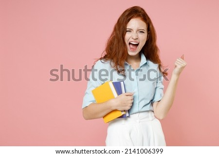 Young happy redhead student woman in blue shirt hold book do winner gesture clench fist celebrate isolated on pastel pink background studio portrait. Education high school university college concept.