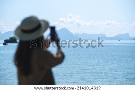Blurred rear view image of a young woman with hat holding and using mobile phone to take photo of the sea