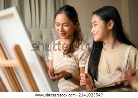 Beautiful woman enjoy painting the artwork on canvas easel with her younger sister in living room.