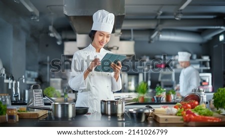 World Famous Restaurant: Asian Female Chef Cooking Delicious and Authentic Food, Uses Digital Tablet Computer While Working in a Modern Professional Kitchen. Preparing gourmet organic Dishes Royalty-Free Stock Photo #2141026799
