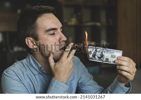 Young brutal businessman lighting cigar with 100 dollar bill as a symbol of wealth and success. The concept of wealth and extravagance. Royalty-Free Stock Photo #2141031267