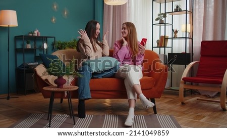 Cheerful girls friends siblings celebrate success win scream rejoices while doing online shopping on smartphone. Happy two female women couple family on sofa at home spend leisure time on mobile phone