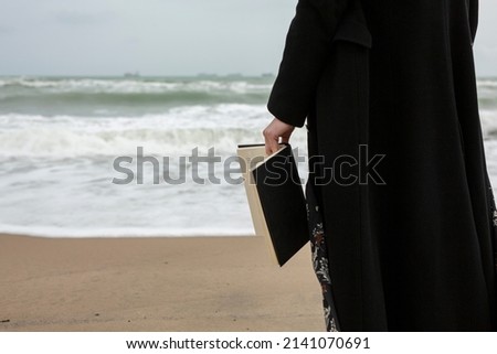 Close-up of open book with black covers held in the hands of a woman on the beach during a cloudy day.