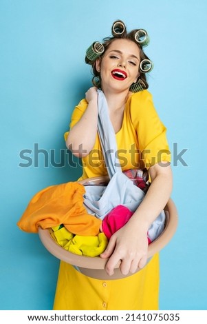 Laundry, cleaning, ironing. Comic portrait of young emotional girl, housewife with curlers on her head isolated on blue studio background. Emotions, female rights, beauty, family, stereotype