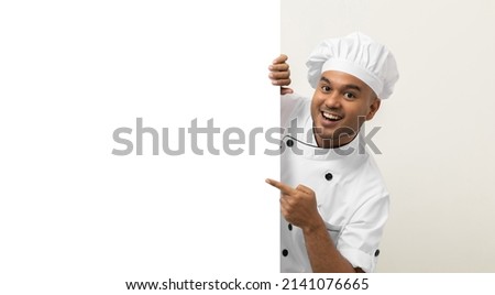 Happy Young handsome asian man chef in uniform looks out from behind an empty whiteboard. Cooking indian man hiding behind big blank billboard for advertising text menu in kitchen and restaurant. Royalty-Free Stock Photo #2141076665