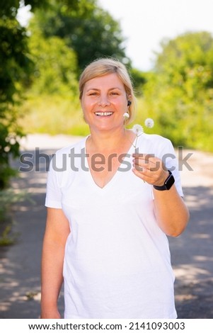 Picture of a happy sporty woman standing on the road in the forest wearing aid and digital watch, resting after jogging and holding the dandelion in one hand.