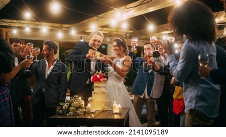 Beautiful Bride and Groom Celebrate Wedding at an Evening Reception Party with Multiethnic Friends. Married Couple Standing at a Dinner Table, Kiss and Cut Wedding Cake. Royalty-Free Stock Photo #2141095189