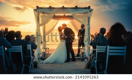 Beautiful Bride and Groom During an Outdoors Wedding Ceremony on an Ocean Beach at Sunset. Perfect Venue for Romantic Couple to Get Married, Exchange Rings, Kiss and Share Celebrations with Friends. Royalty-Free Stock Photo #2141095263