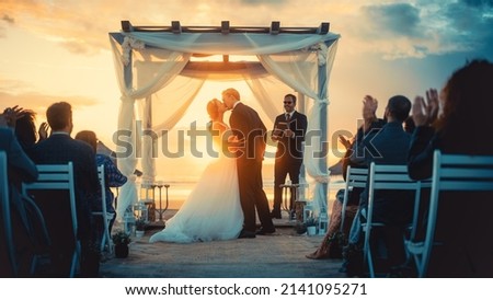 Beautiful Bride and Groom During an Outdoors Wedding Ceremony on an Ocean Beach at Sunset. Perfect Venue for Romantic Couple to Get Married, Exchange Rings, Kiss and Share Celebrations with Friends. Royalty-Free Stock Photo #2141095271