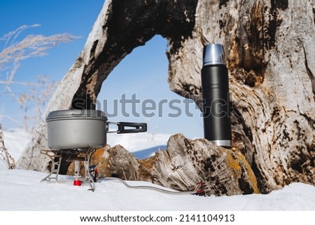 Tourist tableware close-up. A pot of food stands on a gas burner, a black thermos with tea, a winter hike, snow in the forest, cooking in nature. High quality photo