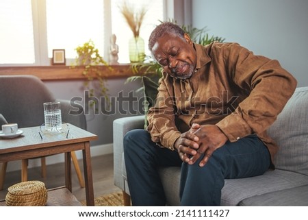 Photo of mature, elderly man sitting on a sofa in the living room at home and touching his knee by the pain during the day. Mature man massaging his painful knee.