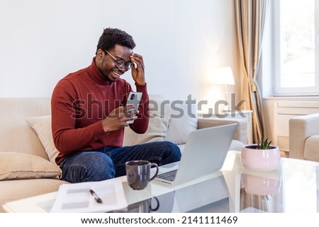 Excited Black African American Man Having a Video Call on Smartphone while Sitting on a Sofa in Living Room. Happy Man Smiling at Home and Talking to His Friends and Family Over the Internet.



