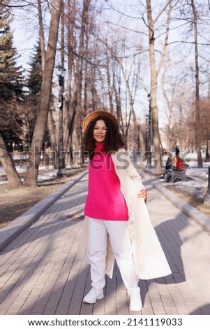 Happy curly brunette girl in beige hat and sun glasses smiling outdoors. Young woman happy walking in street. Pink sweater, beige coat, beige hat. Sun in city. Fashionable asian girl with frizzly hair