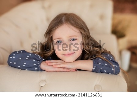  Inspired little girl with big eyes and curly hair. Beautiful young girl rest on couch and looking at camera. Childhood concept. Dreaming child. Smiling little girl sitting on a couch in living room.