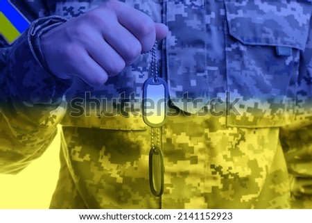 Closeup view of defender in camouflage uniform holding military ID tags, toned in colors of Ukrainian flag