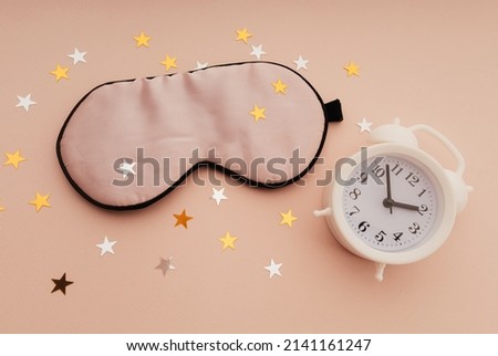 Concept eye protection from light for good sleep. Sleeping mask, white alarm clock and paper stars on pink background.