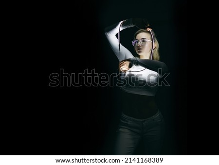 The portrait of beautiful woman in white clothes, top, shiny shimmer makeup and stars with led stripes, neon light on black background. Futuristic style girl in glasses, sadly emotions. Future.