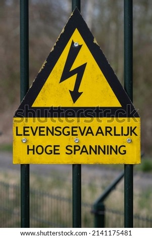 Yellow and black triangular sign with lightning bolt and Dutch text 'Voltage life-threatening' or 'electricity voltage dangerous', mounted on a fence. Vertical image