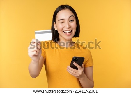 Joyful asian girl smiling, showing credit card and smartphone, recommending mobile phone banking, standing against yellow background
