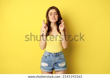 Hopeful young beautiful woman praying, believe, hoping to receive smth, cross fingers for good luck, anticipating, standing over yellow background