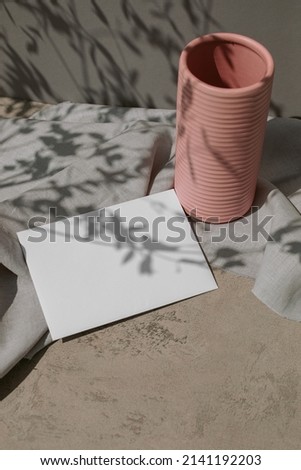Empty white paper mock up, linen cloth, modern ceramic vase, leaves shadows on surface. Still life. 