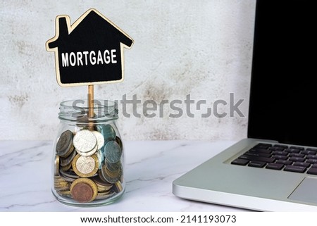Mortgage text on wooden house model with jar full of coins and notebook on white table.