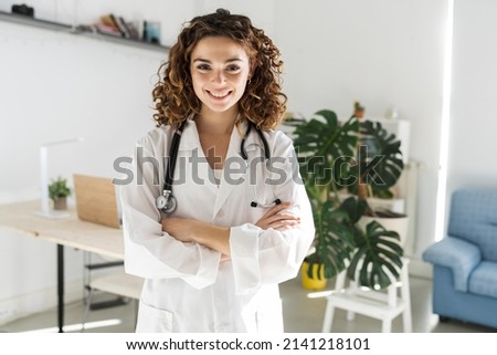 Happy young woman doctor with curly hair wears white medical gown and stethoscope looking at camera. Smiling female doctor, general practitioner consulting patient online. Close up head shot portrait Royalty-Free Stock Photo #2141218101